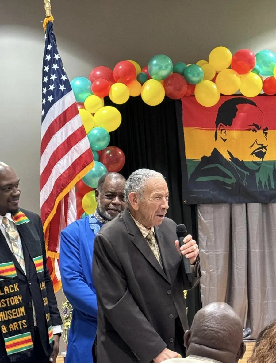 An older gentleman in a dark suit speaks into a microphone beside an American flag at a Dr. Martin Luther King Jr. celebration